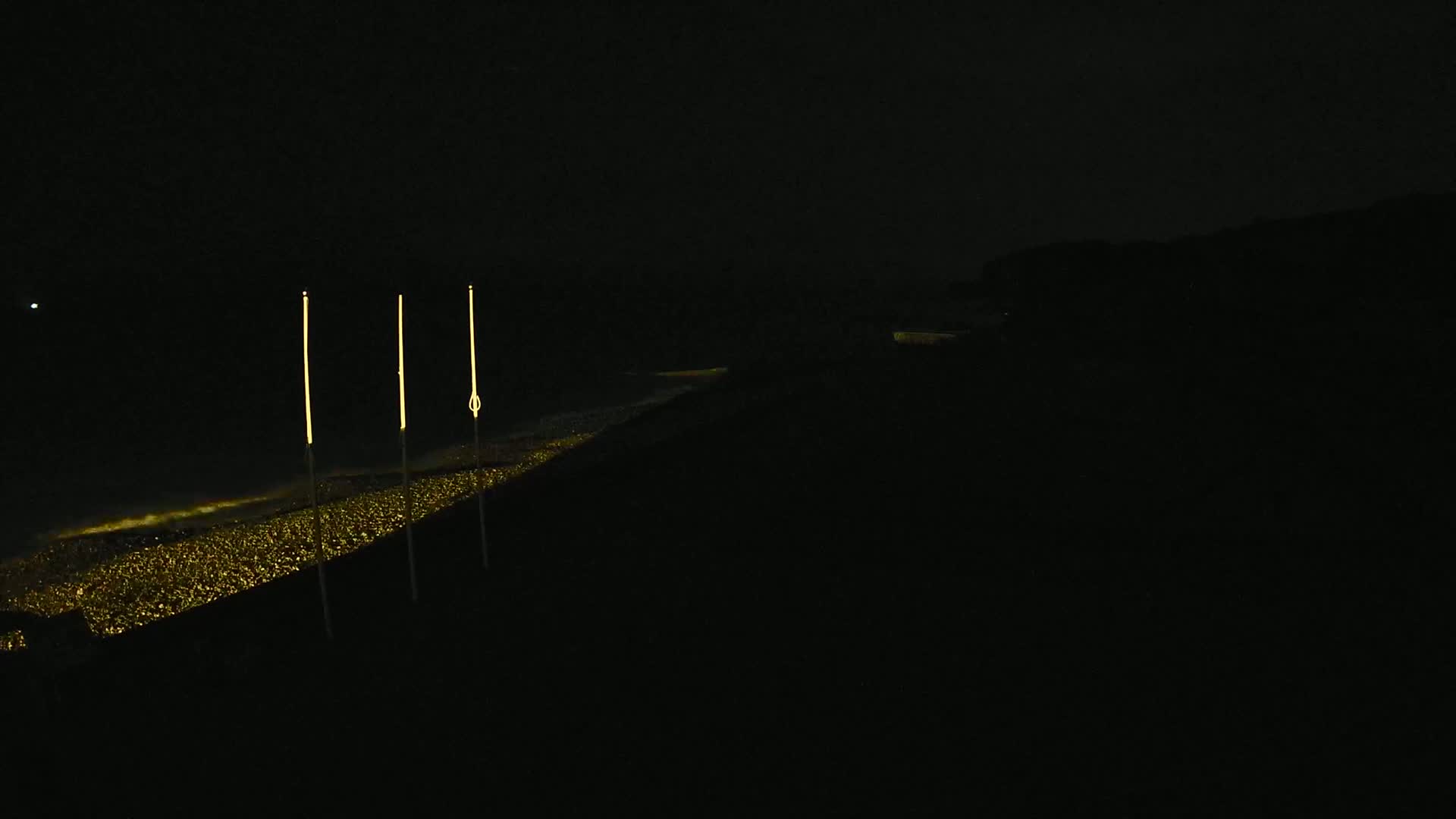 At night, three tall, thin lights stand illuminated above a long, low expanse of glittering gold, reflecting in a dark body of water.