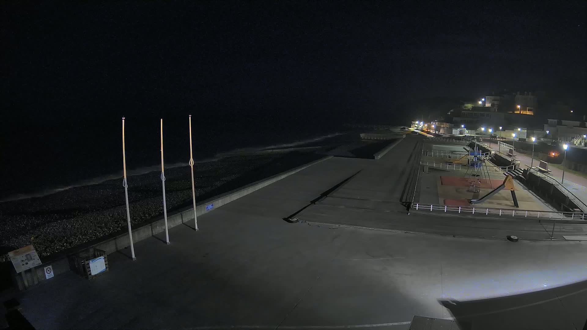 A nighttime view of a paved seaside area with a playground, a low wall separating it from a dark, rocky beach with small waves, and buildings illuminated in the background under a clear, dark sky.