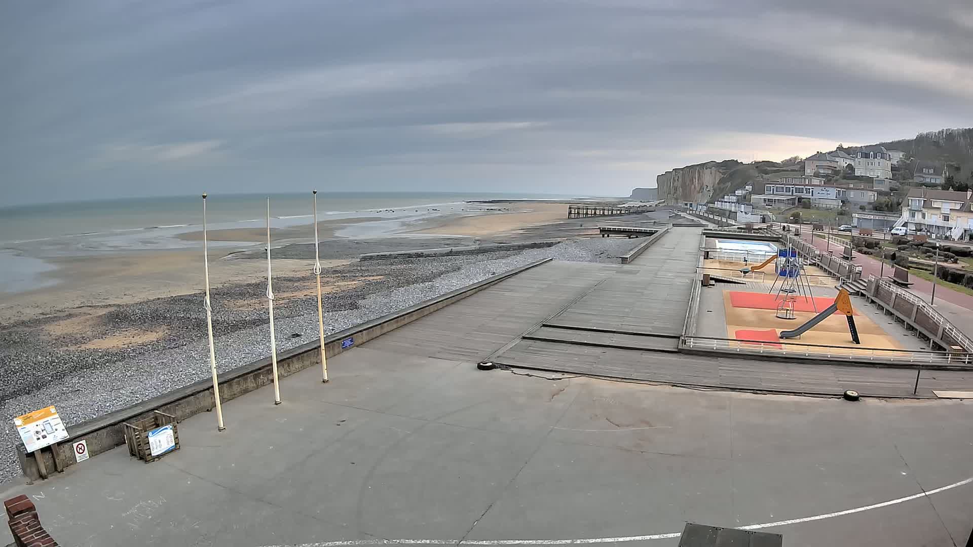 A beach with a playground and a paved walkway overlooks a calm ocean under a cloudy sky.