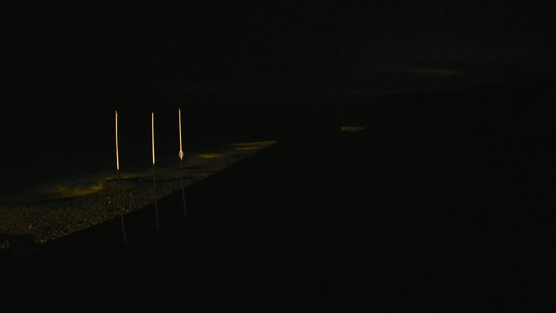 Three tall, slender lights illuminate a dark, granular surface at night, reflecting faintly in the water below.