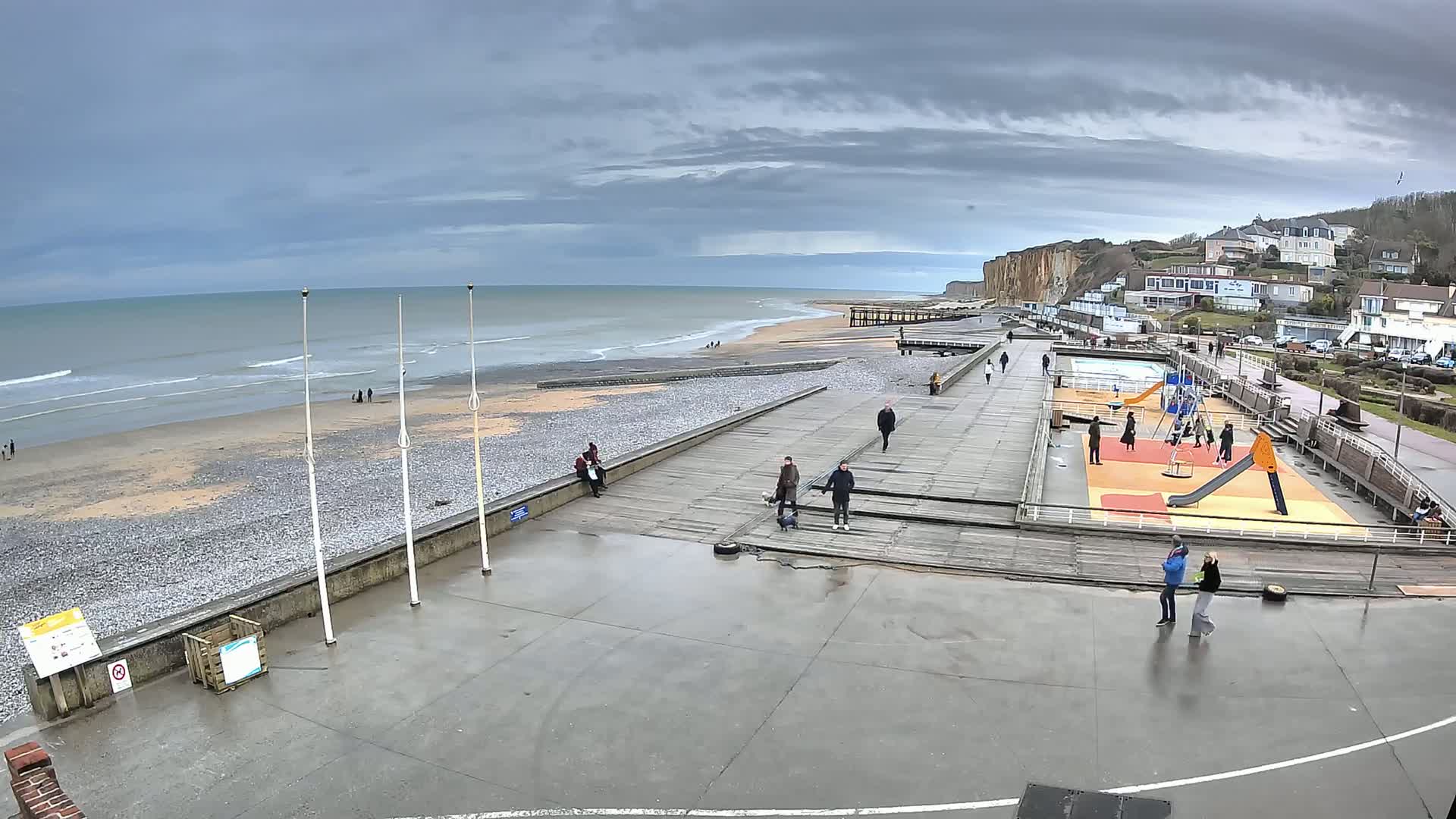 A wet, overcast day at a pebble beach with a boardwalk, playground, and a few people scattered around.