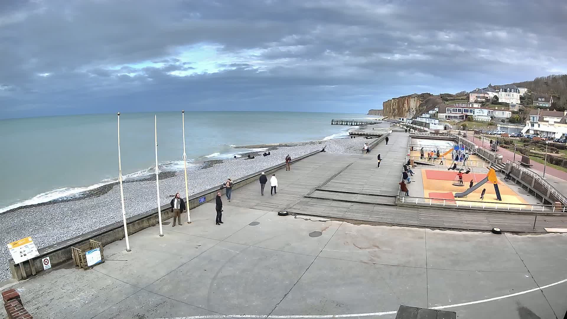 A seaside promenade with a playground, a pebble beach, and a pier under a cloudy sky.