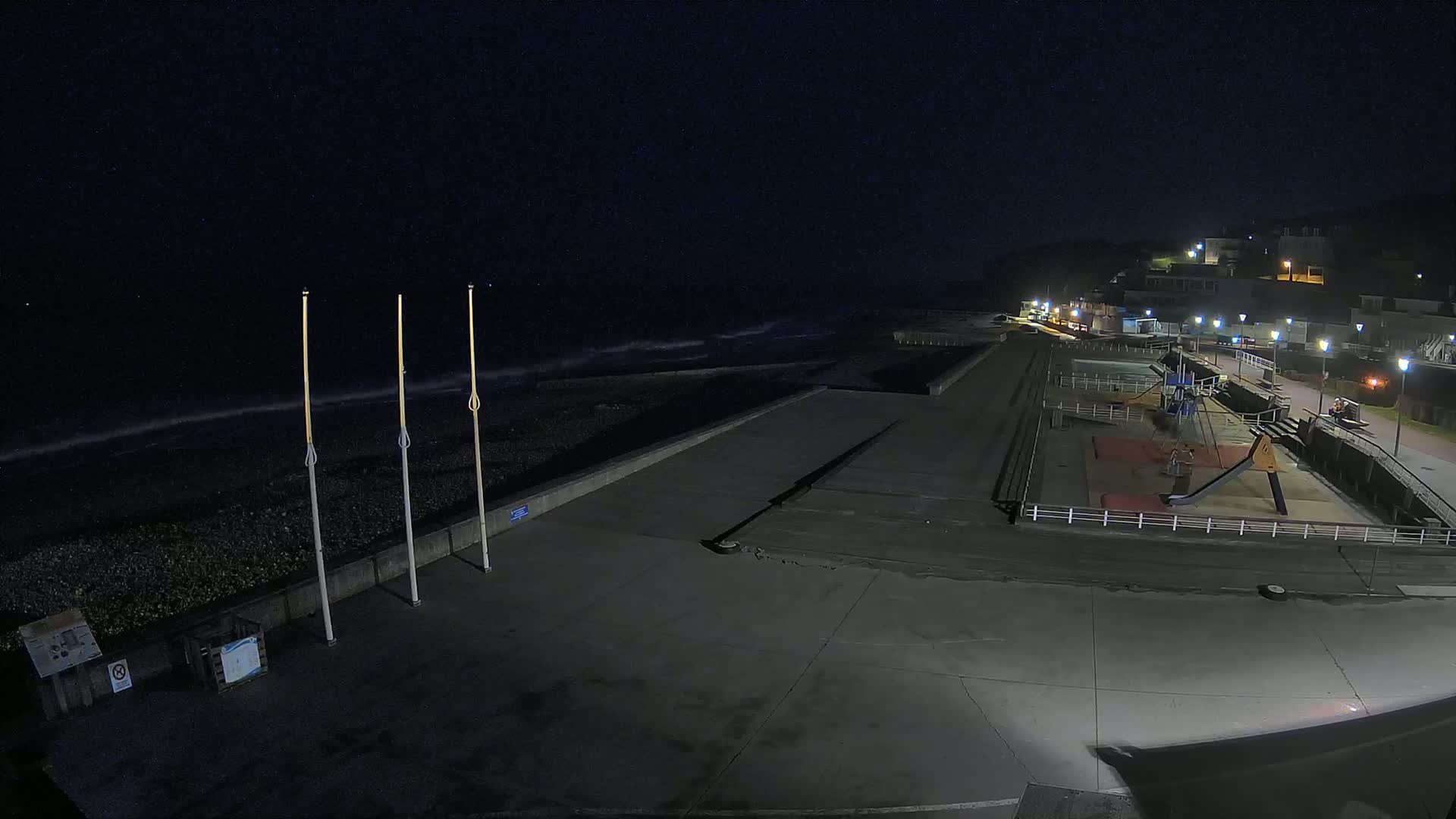 A nighttime view of a mostly empty paved beachfront area with a playground and a rocky shoreline under a dark, clear sky.