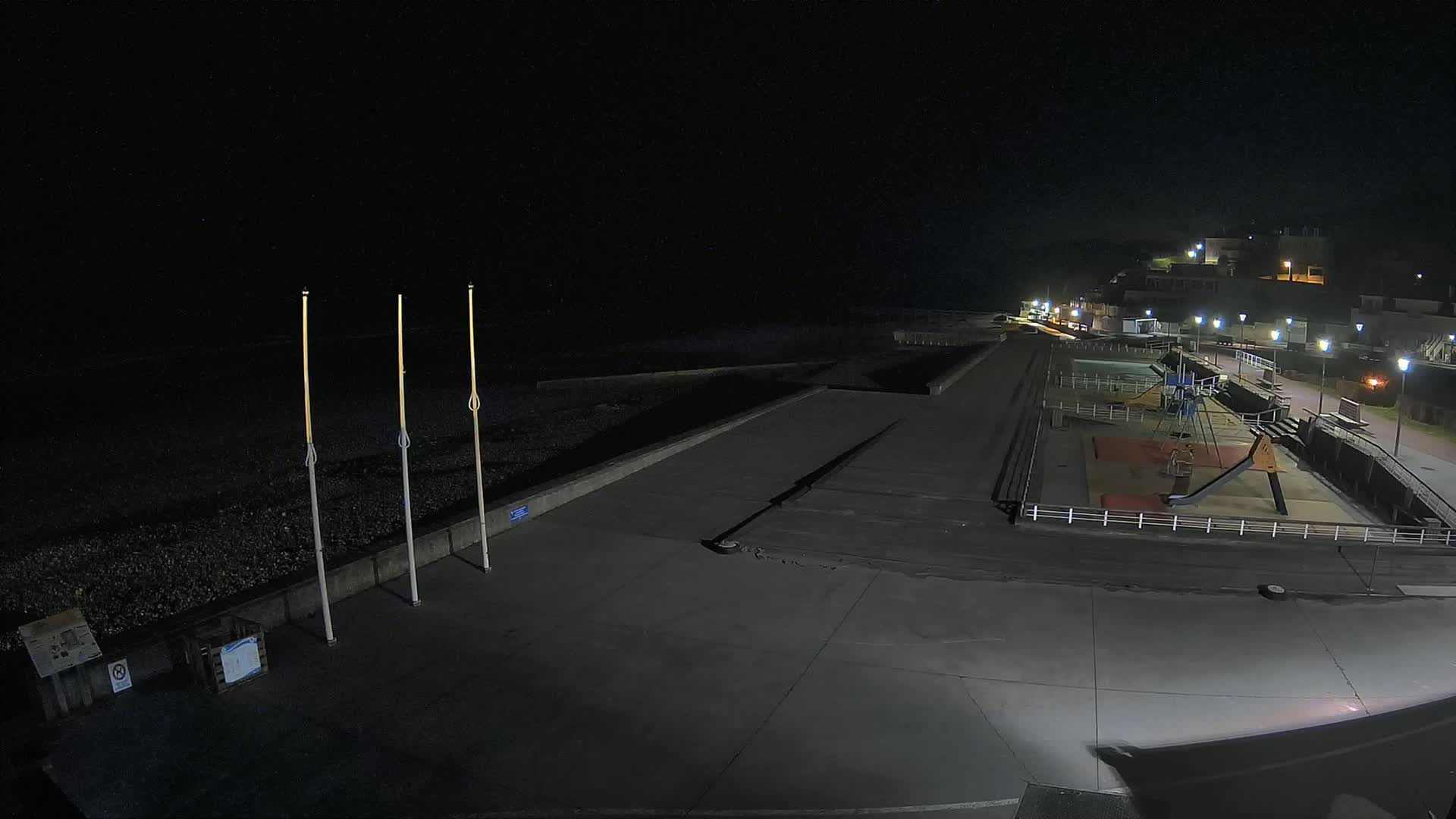 At night, a mostly empty paved beachfront area with a playground and a low seawall is illuminated by streetlights.