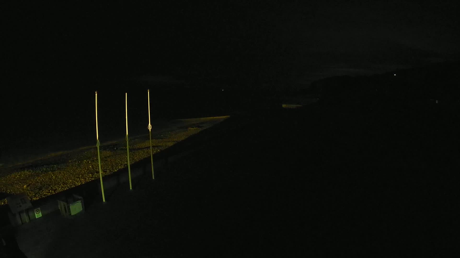 Three tall, thin lights stand illuminated on a dark pebble beach at night.