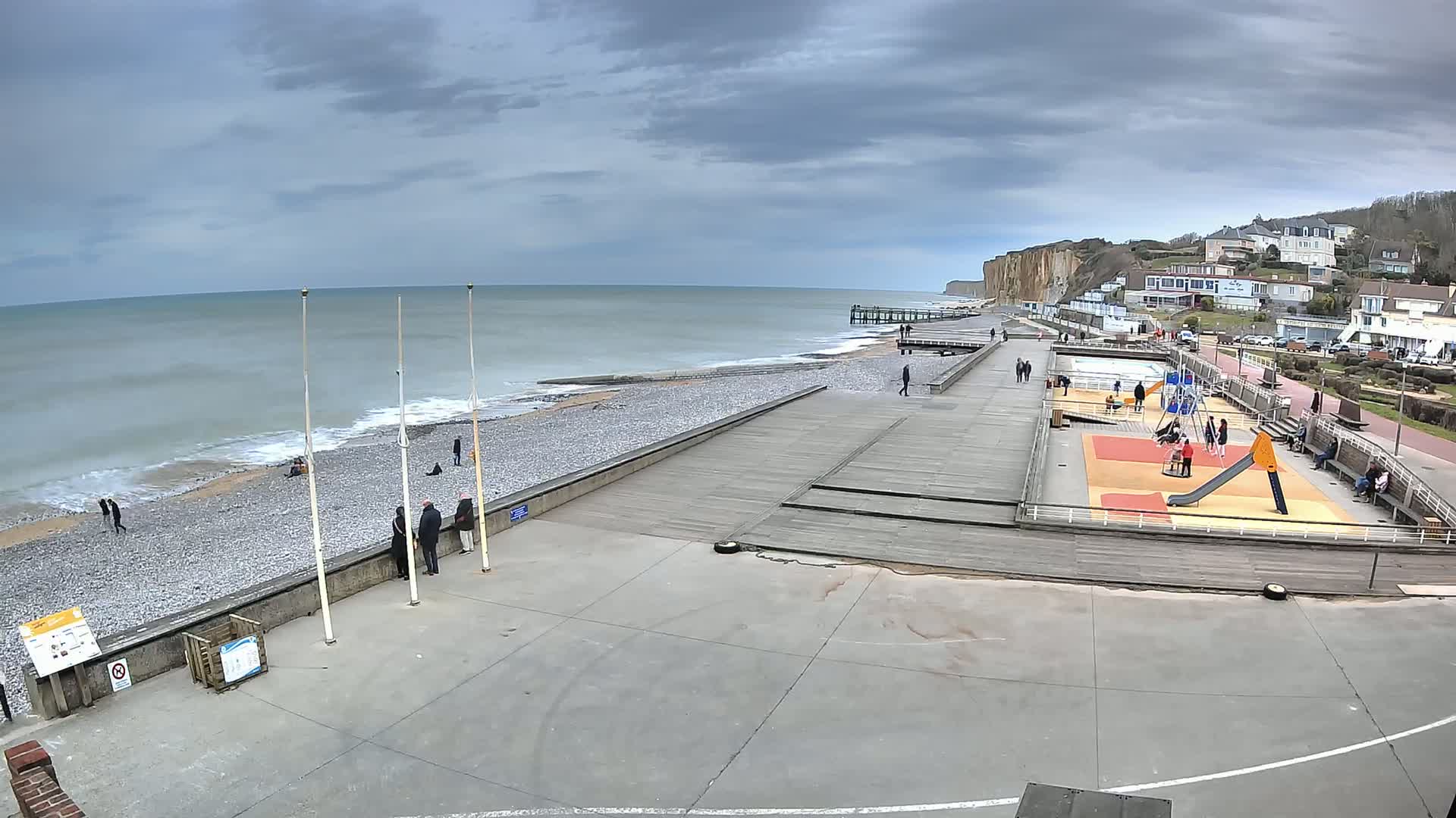 A pebble beach with a wooden boardwalk and playground is seen under an overcast sky, with a calm sea and high cliffs in the background.