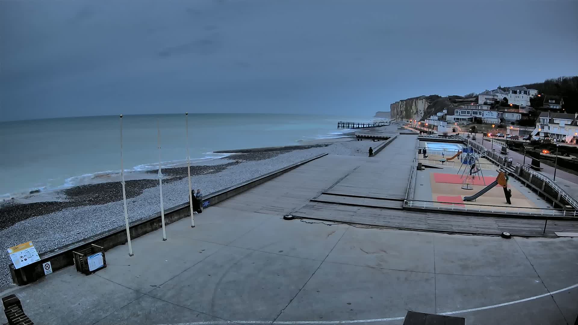 A mostly empty beach and boardwalk with a playground and small pool, under an overcast sky, stretches along a rocky shoreline next to white cliffs and houses.