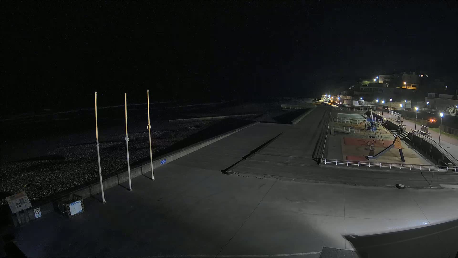 A nighttime, illuminated view of an empty seaside promenade with a playground and a dark, calm ocean in the background.