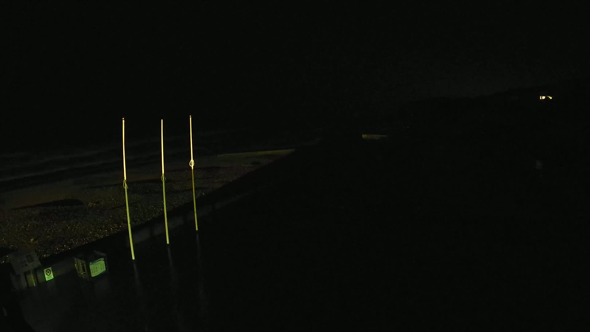 Three tall, thin, illuminated posts stand on a dark beach at night next to dark water.