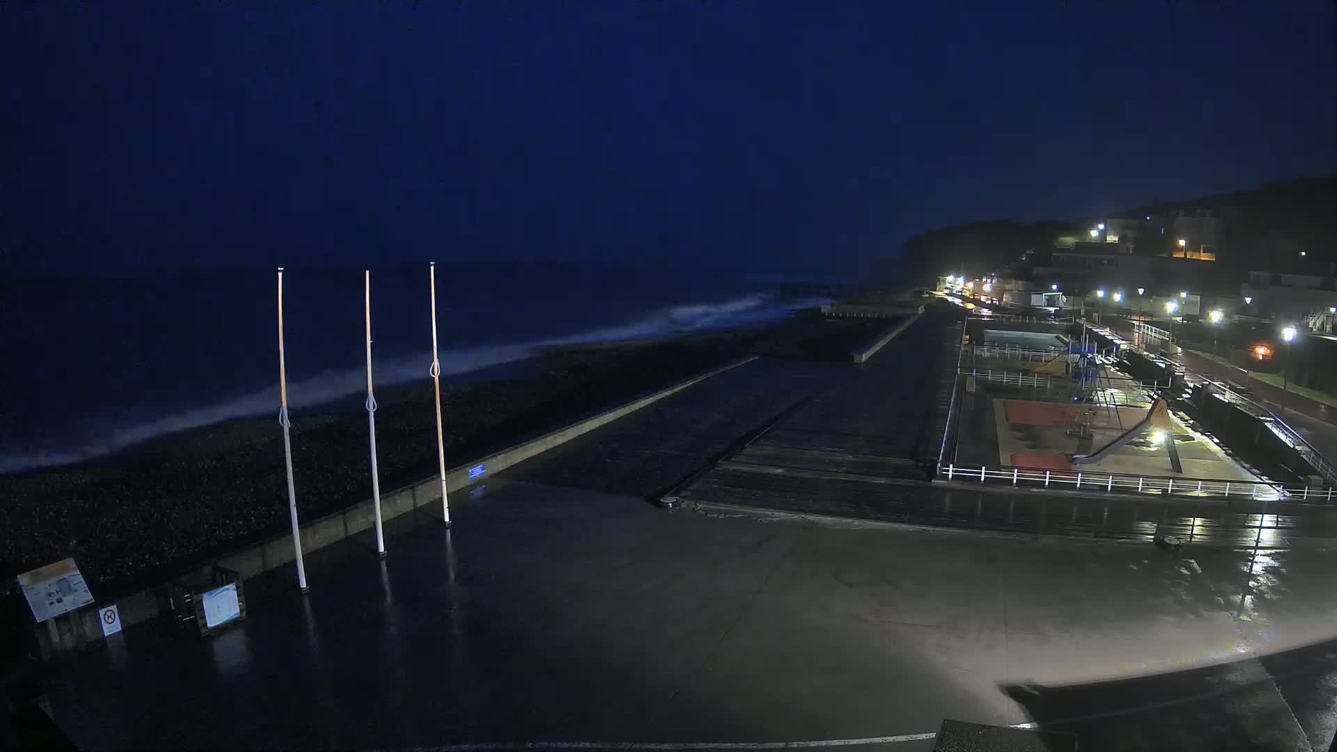 A nighttime seaside scene shows dark, wavy water crashing against a shoreline with a playground and pathway illuminated by streetlights under a dark, overcast sky.