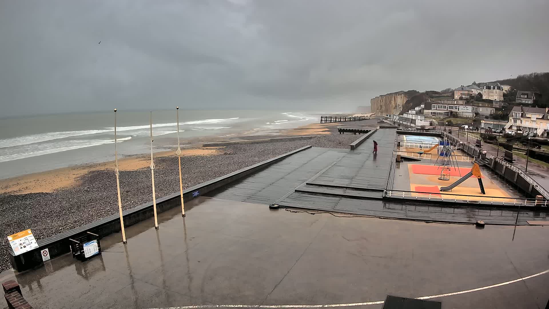 A wet, paved beachfront area with a playground and a view of a stormy gray sea and white cliffs in the distance.