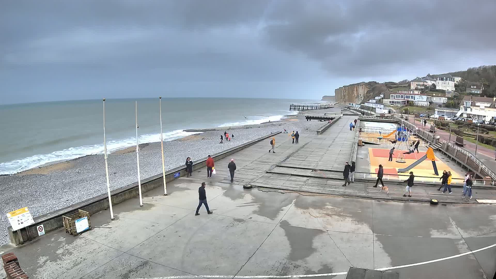 A pebble beach with a wooden boardwalk and playground overlooks a gray, choppy sea under a cloudy sky.
