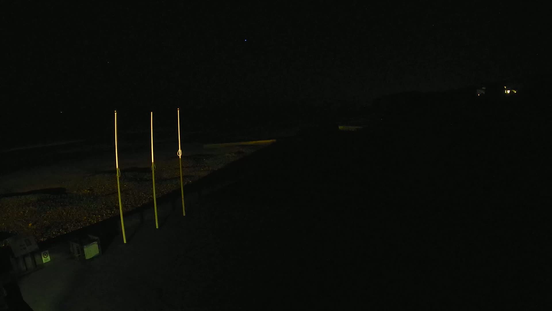 Three tall, slender, illuminated posts stand on a dark, pebble-covered surface at night under a clear, starlit sky.
