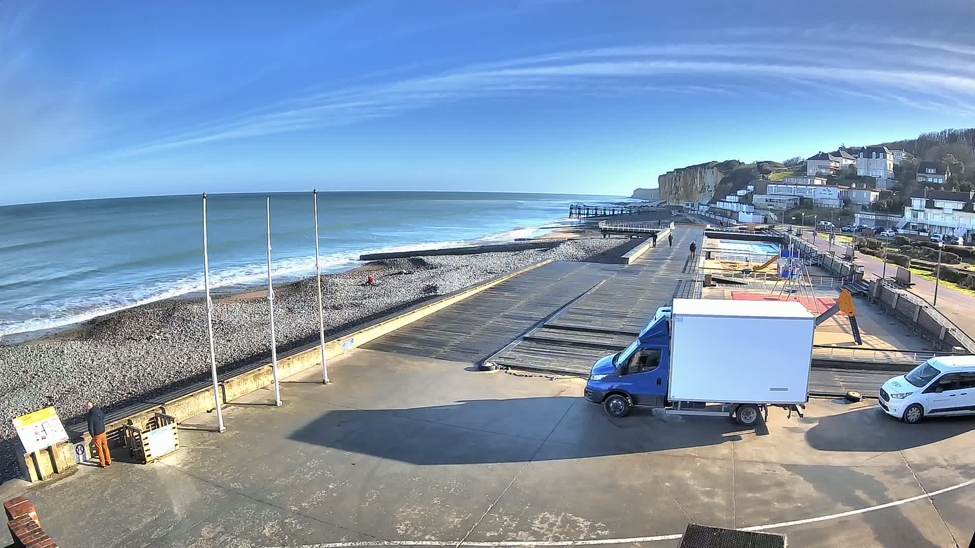 A seaside scene on a sunny day shows a pebble beach, a wooden boardwalk, a small playground, and parked delivery vehicles next to a low cliff and buildings.
