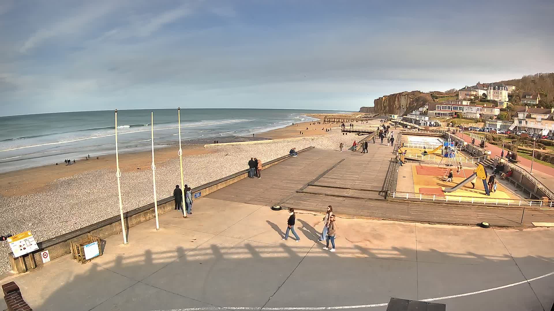A mostly sunny day at a beach with a pebbled shore, wooden boardwalk, playground, and buildings on a cliff in the background.