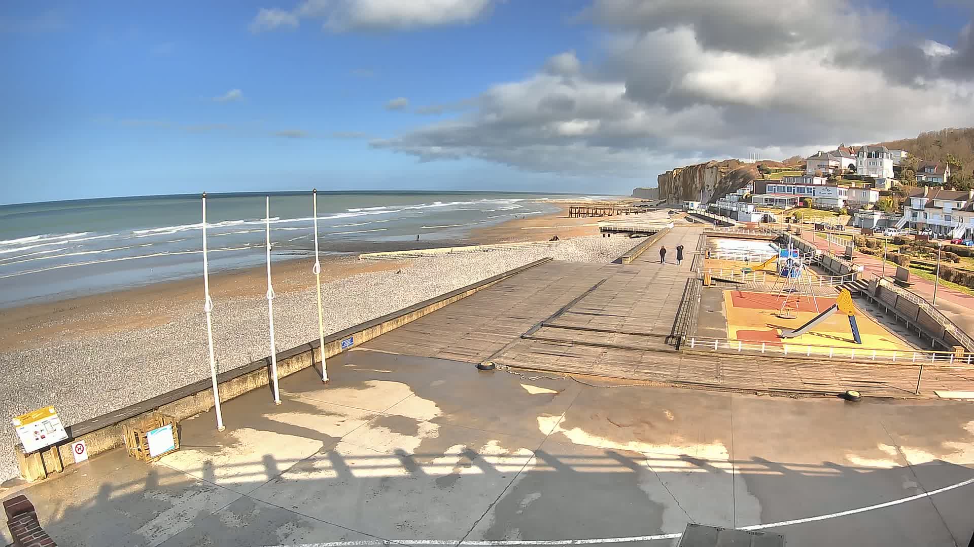 A pebble beach with a wooden boardwalk, playground, and buildings along a cliffside under a partly cloudy sky.