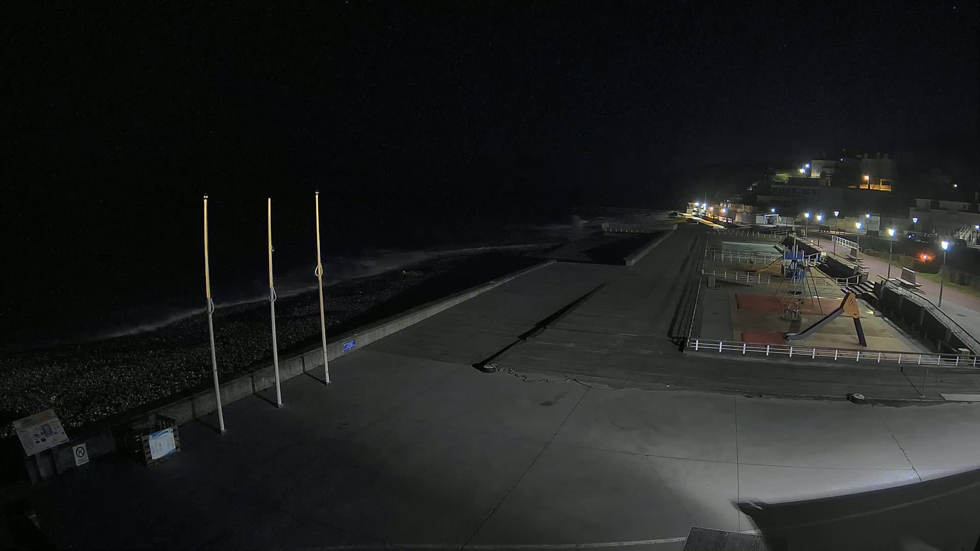 A nighttime view of a mostly empty seaside promenade with a small playground and a dark, wavy ocean in the background under a clear, starlit sky.