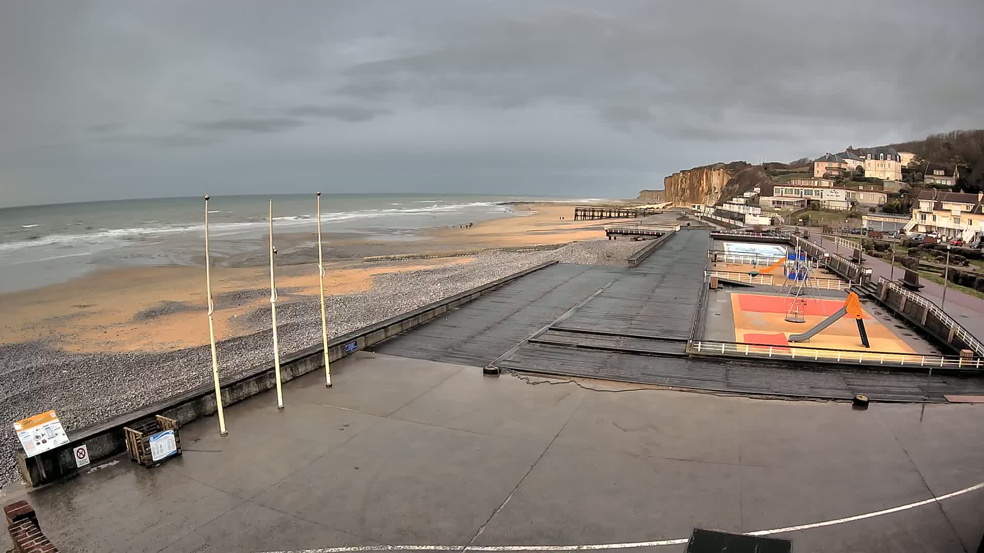 A beach with a playground and wooden boardwalk is shown under an overcast sky, with waves gently rolling onto the shore.
