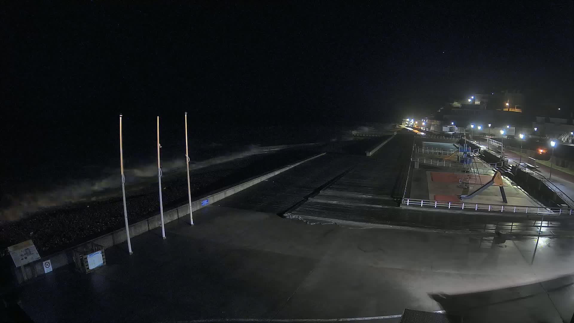 A nighttime coastal scene shows a playground and a paved area next to a seawall with rough waves crashing against it under a dark sky.
