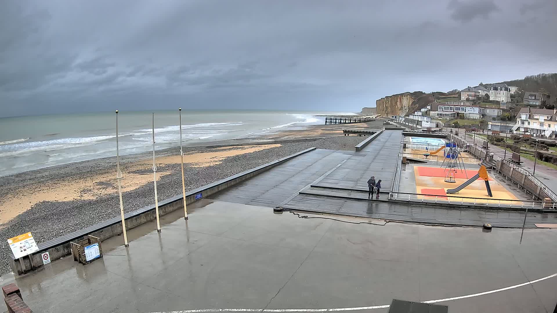 A wet, overcast day at a beach with a playground and a boardwalk next to a rocky shoreline and high cliffs, showing a small group of people walking.
