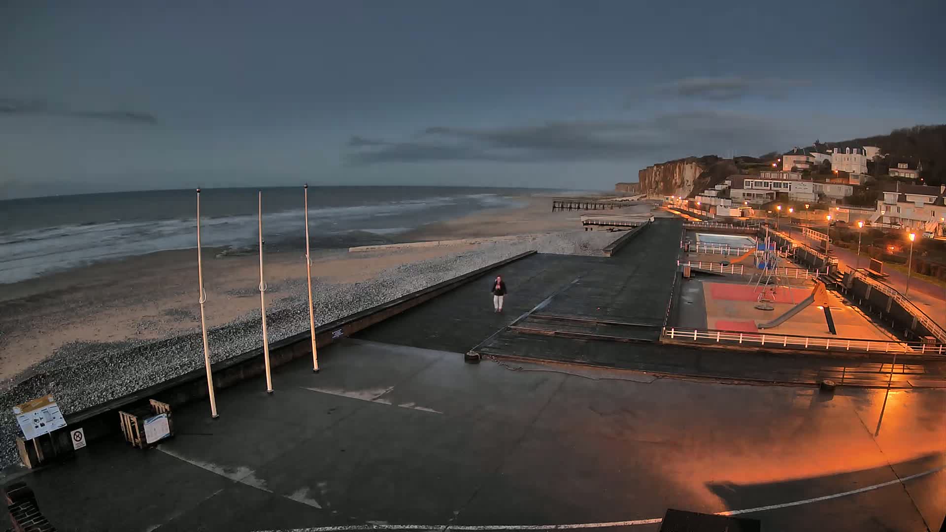 A person walks on a paved beachfront area near a playground and a small pool, with a rocky beach and choppy ocean under a dark, cloudy sky.