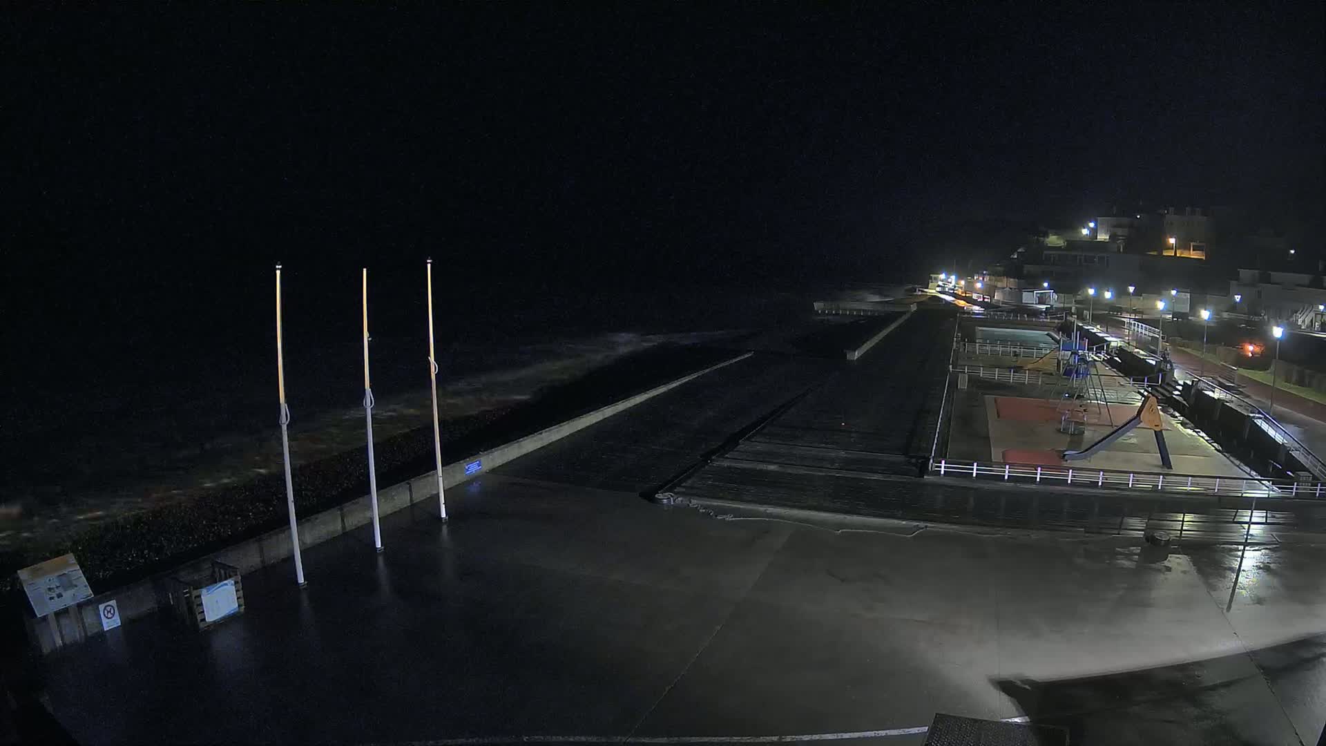 A nighttime view of a wet, empty seaside promenade with a playground and crashing waves.