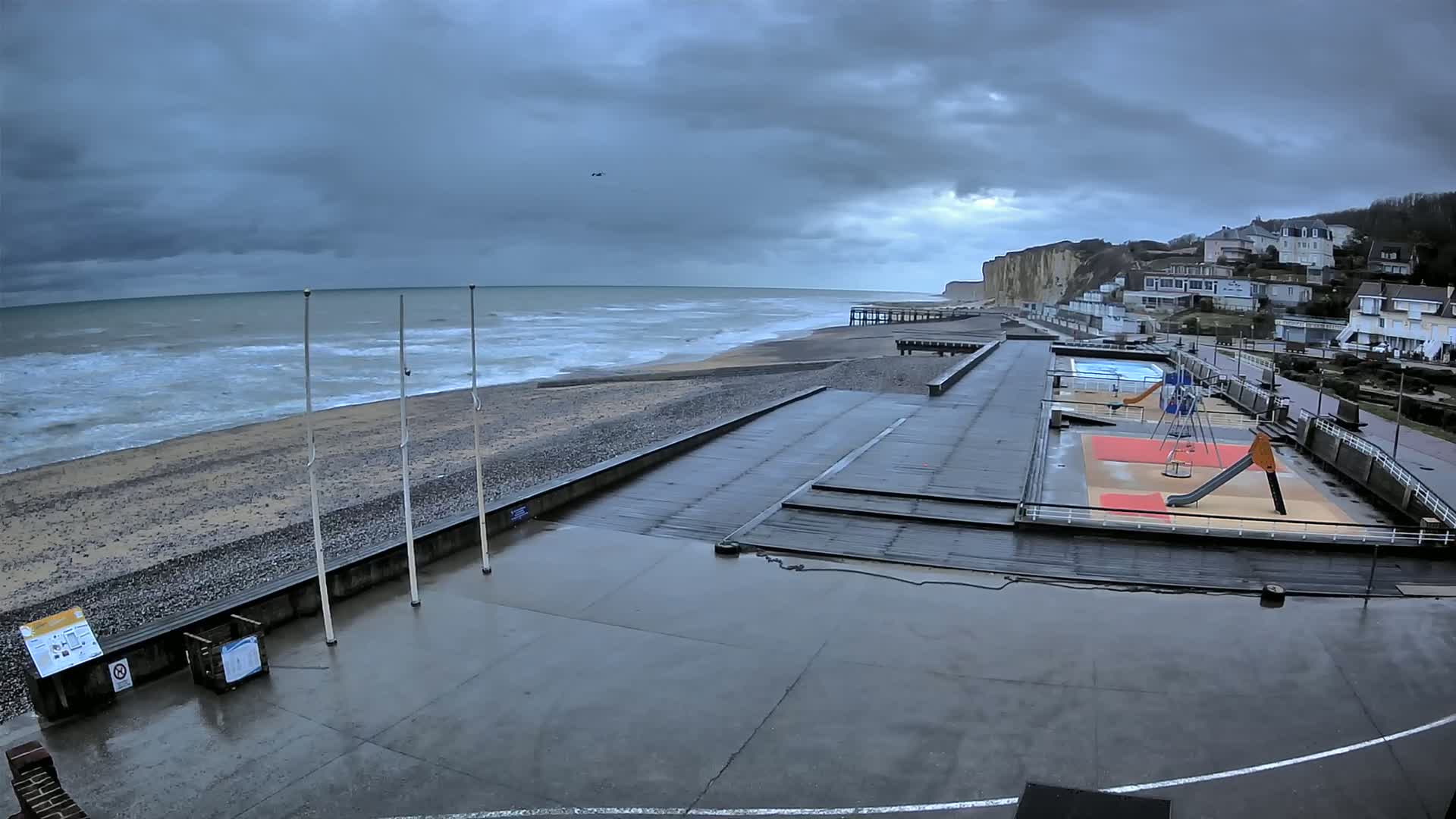 A wet, overcast day at a beach with a playground and a small pier, overlooking a rocky shore and dramatic cliffs.