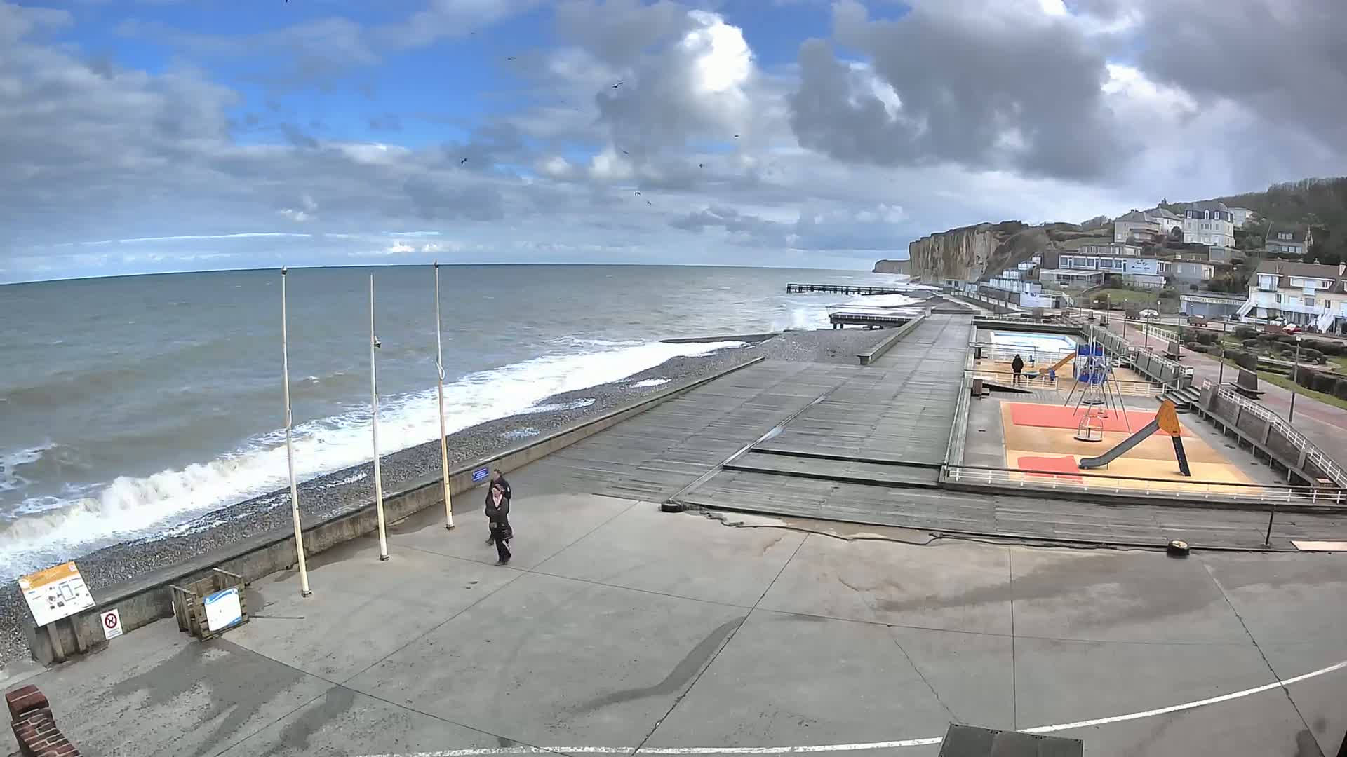A seaside boardwalk with a playground and a couple walking, under a partly cloudy sky with a choppy sea.