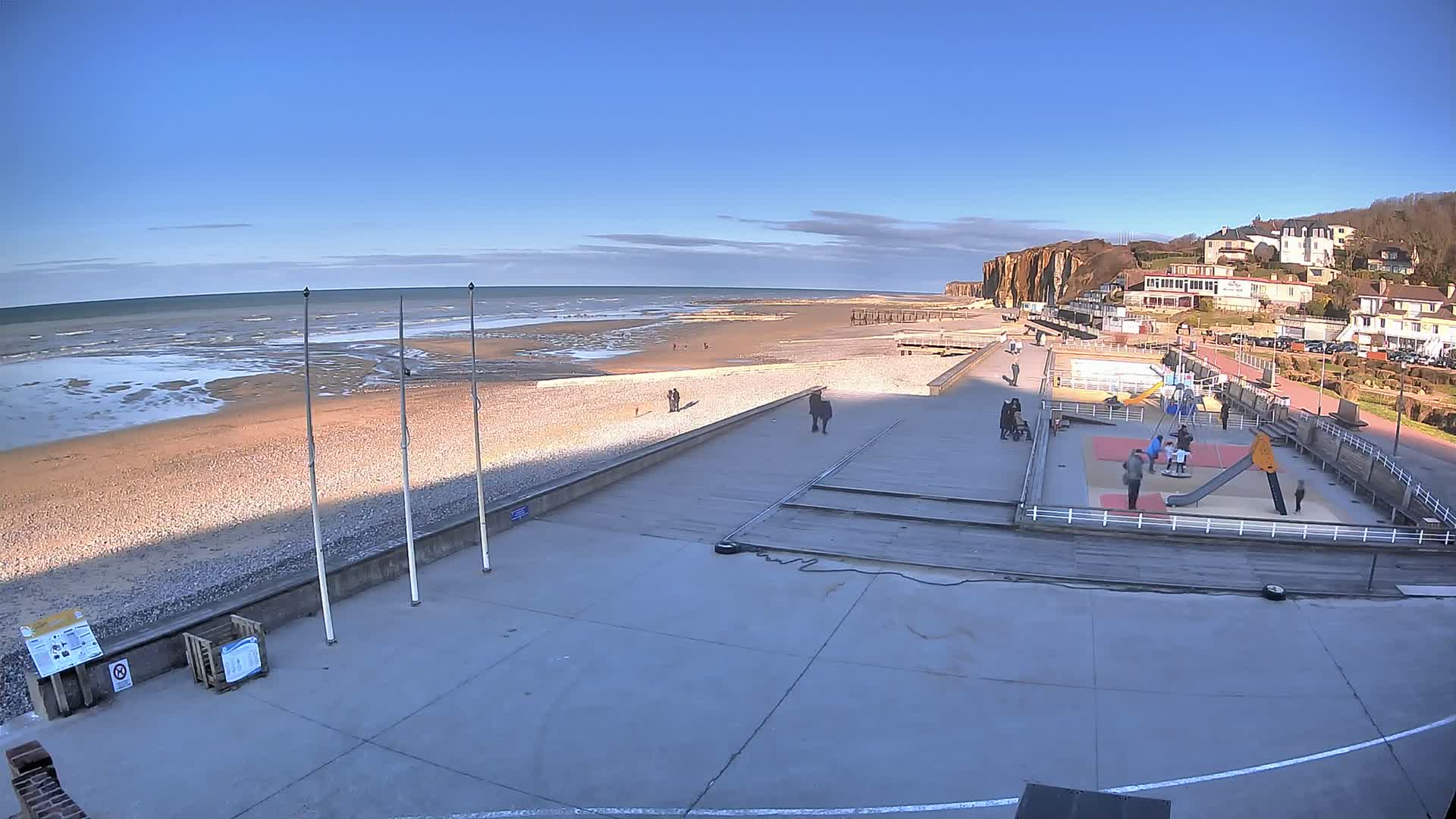 A beach with a playground and a few people is visible under a clear blue sky, with calm waves lapping at the shore.