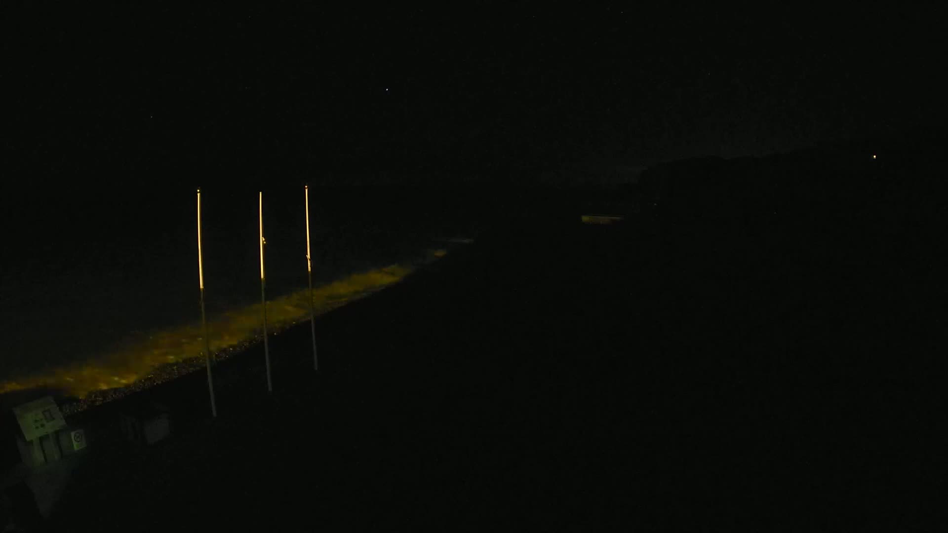 Three tall, slender lights illuminate a dark shoreline at night, their reflections visible in the water.