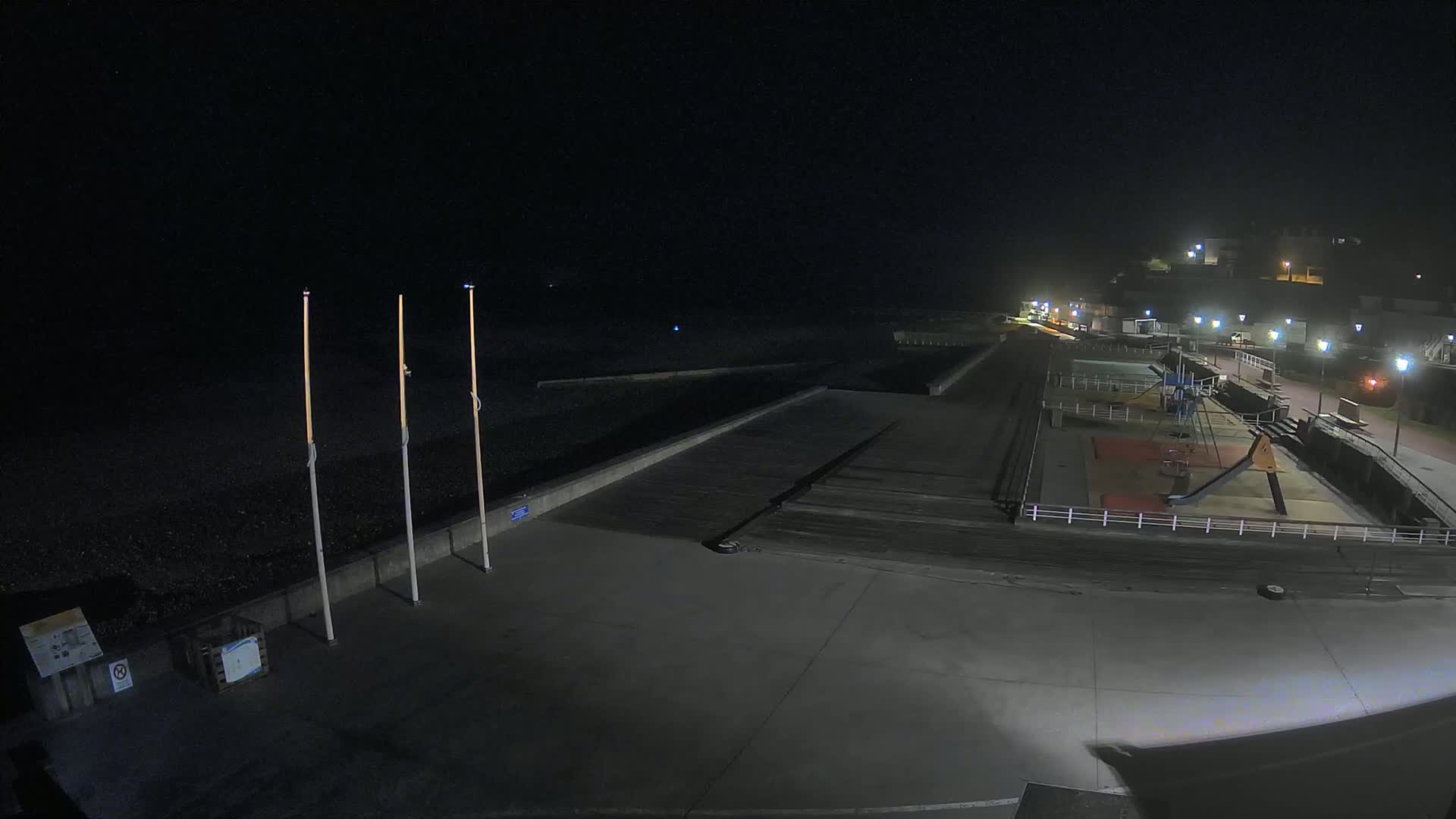 A nighttime, dimly lit view of a mostly empty beachside area with a playground and a dark, pebble beach.