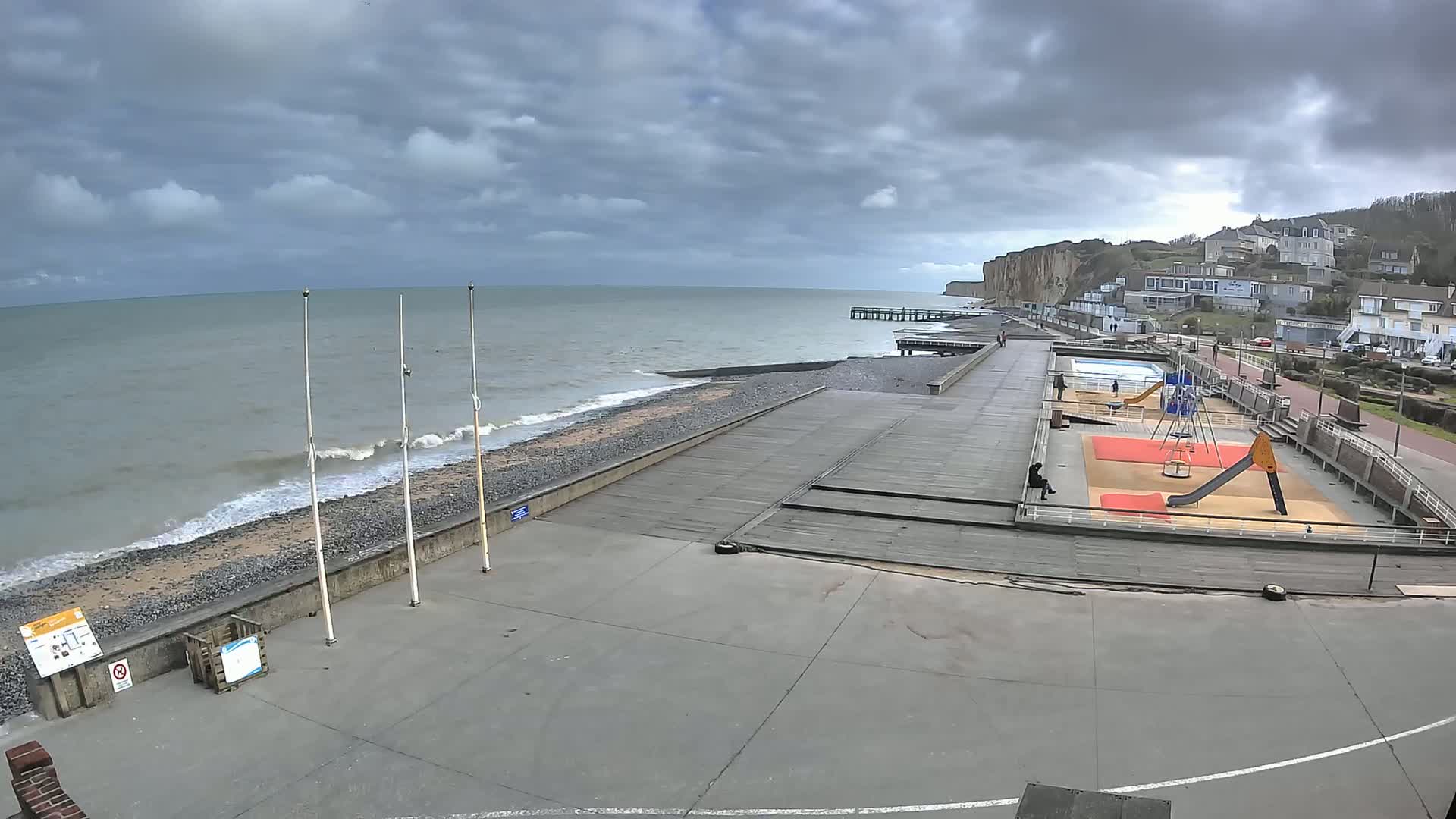 A seaside promenade with a playground and a small pier under a cloudy sky, with gentle waves lapping a pebble beach.