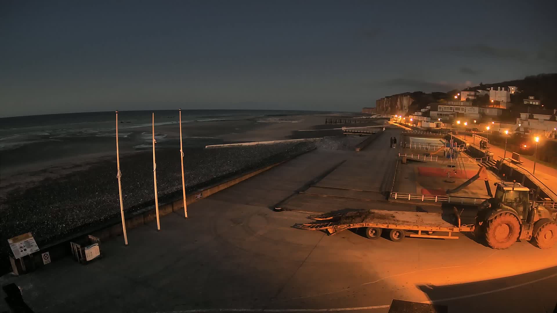 A tractor and trailer sit on a paved area next to a dark, pebble beach under a twilight sky, with a seaside town visible in the background.