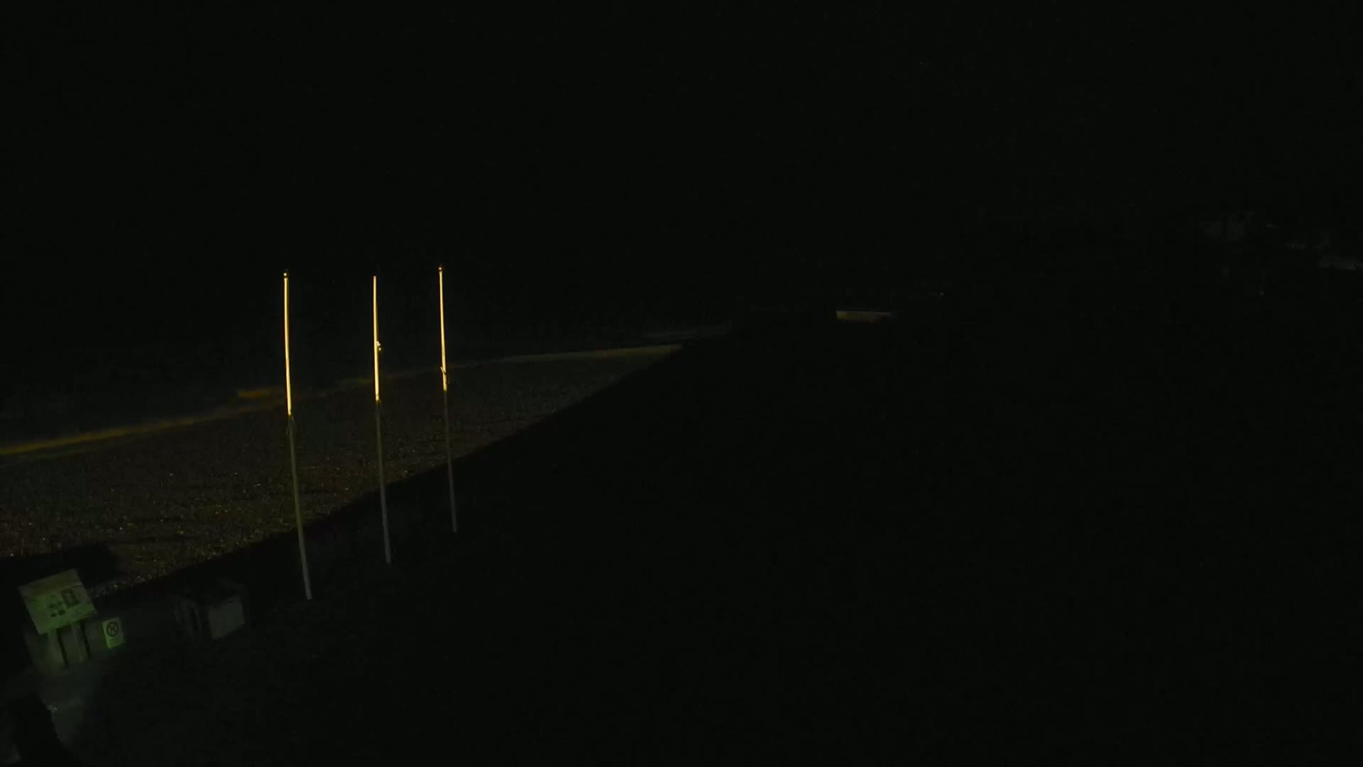 Three tall, slender lights illuminate a dark, pebble-covered beach at night.