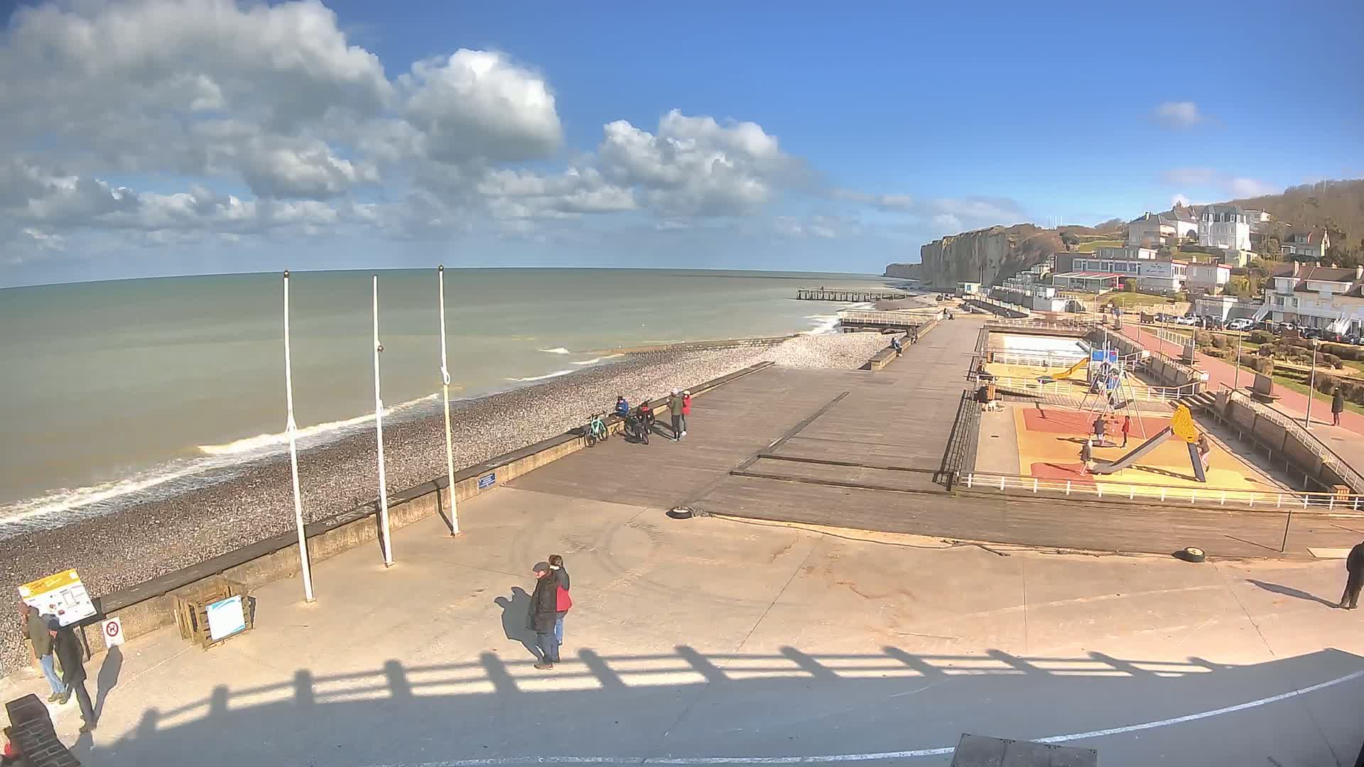 A mostly sunny day at a seaside promenade features a pebbled beach, a wooden boardwalk, a playground, and a few people scattered about.