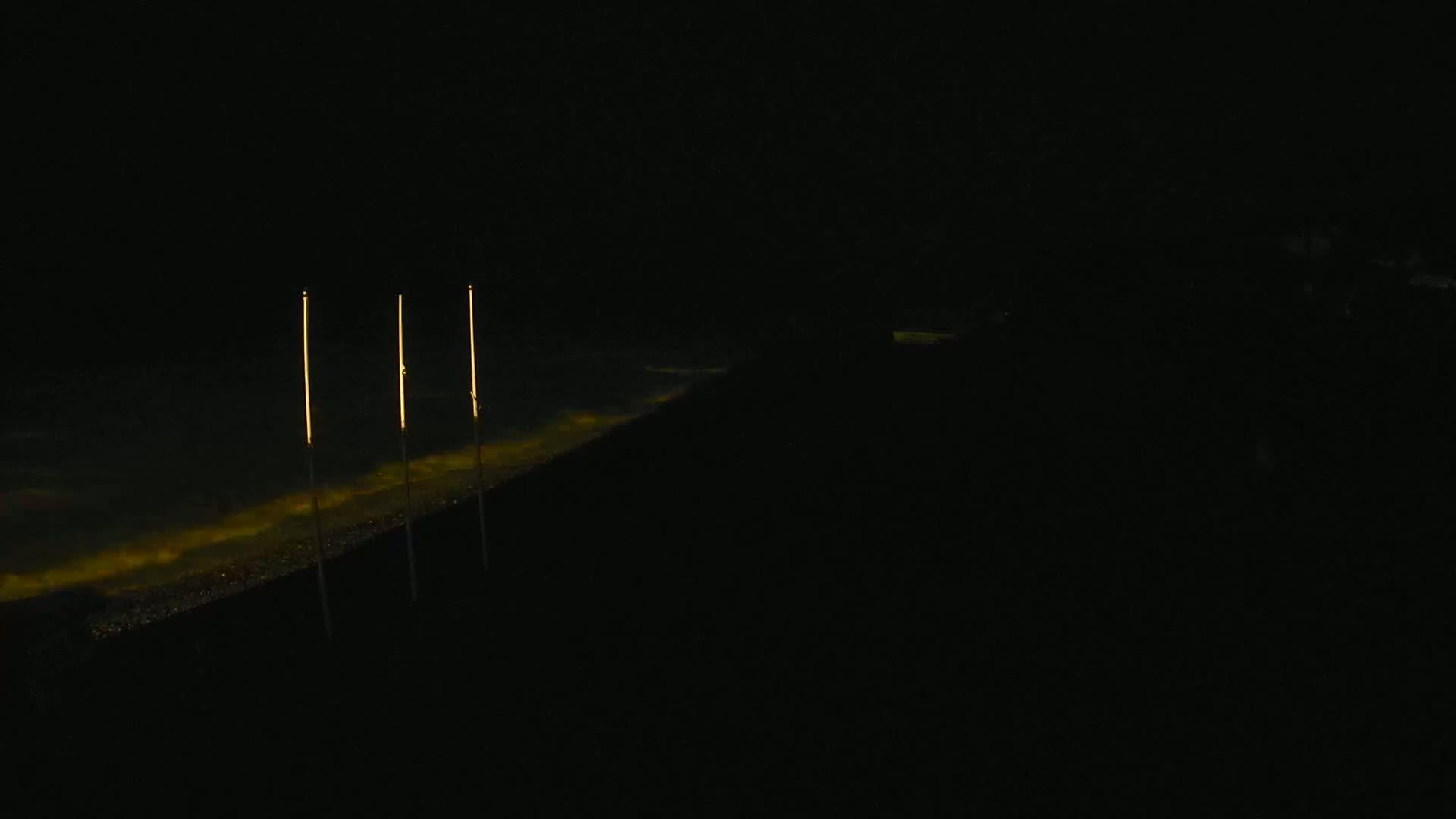 Three tall, thin, illuminated poles stand along a dark, water's edge at night.