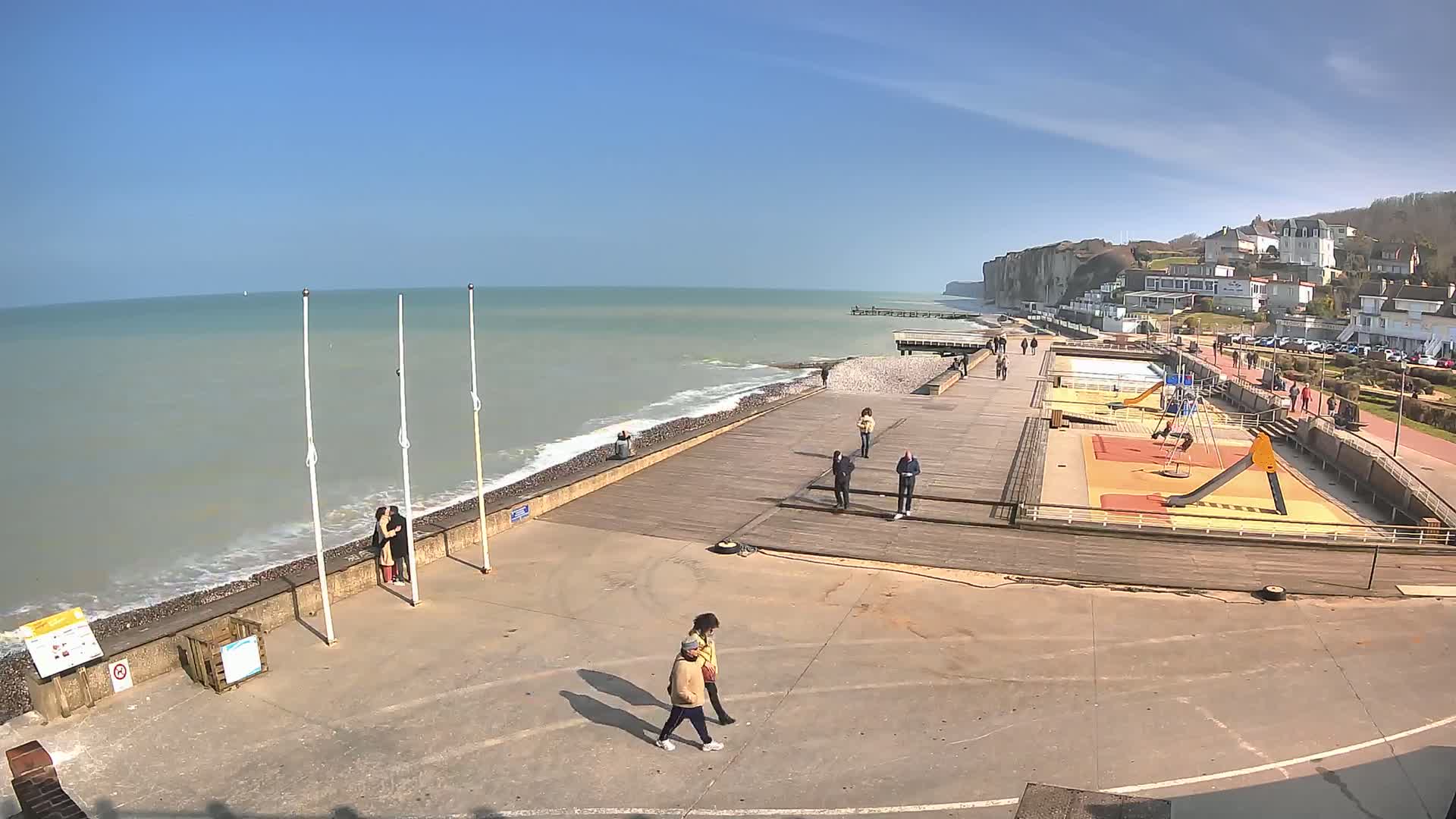 A boardwalk overlooks a rocky beach and ocean with small waves under a clear blue sky; a playground and several people are visible on the boardwalk.