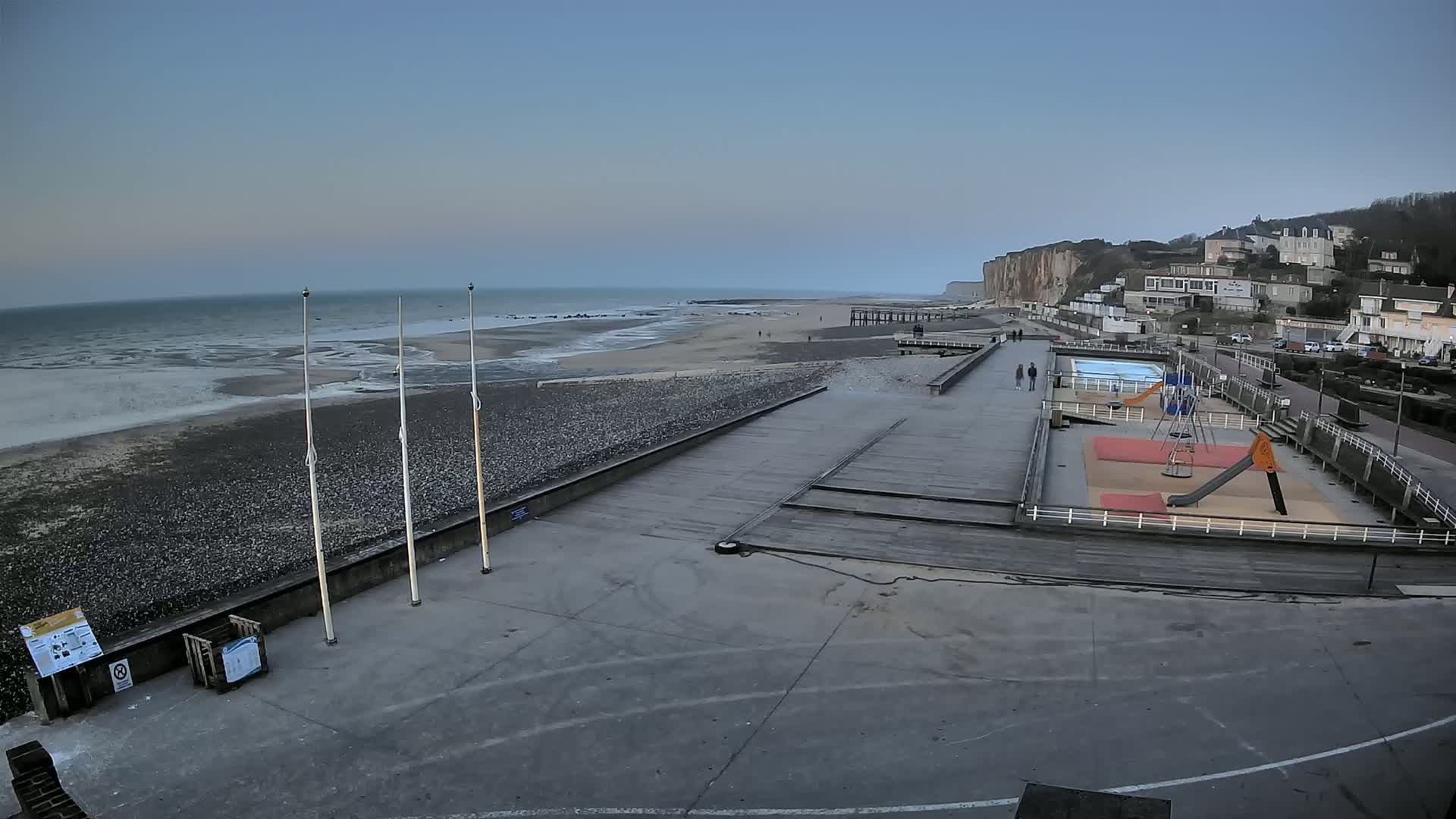 A mostly empty beach with a playground and a few people visible, under a clear, pale sky.