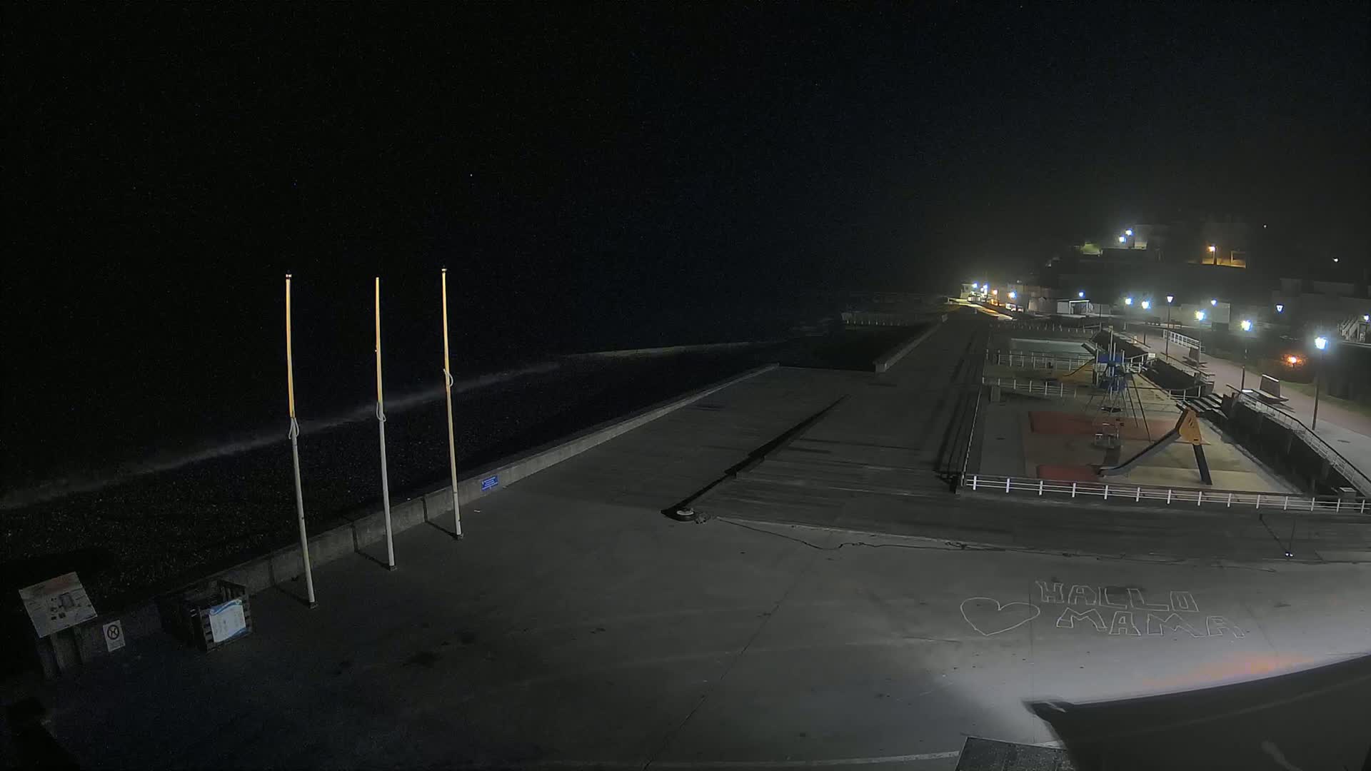 A nighttime, seaside view shows a mostly empty paved area next to a dark ocean with small waves, featuring a playground and some streetlights.