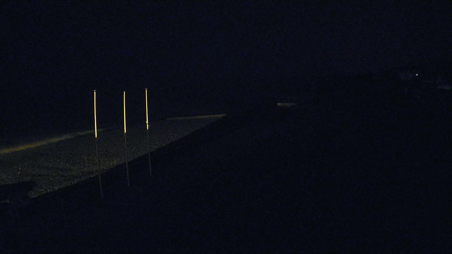 Three tall, thin, illuminated poles stand on a dark, pebbled beach at night.