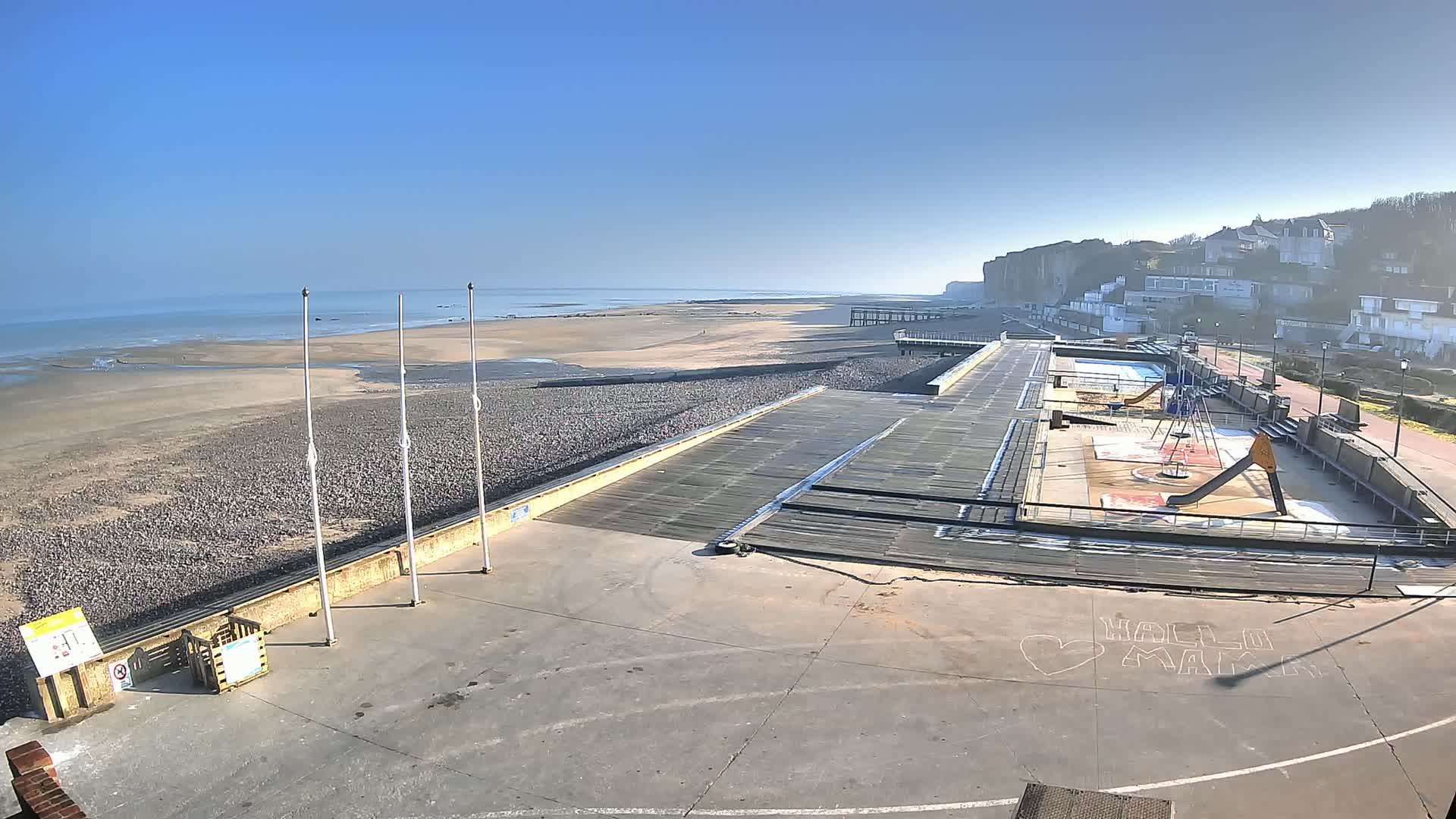 A mostly empty beach and boardwalk with a playground and a calm ocean under a clear blue sky.