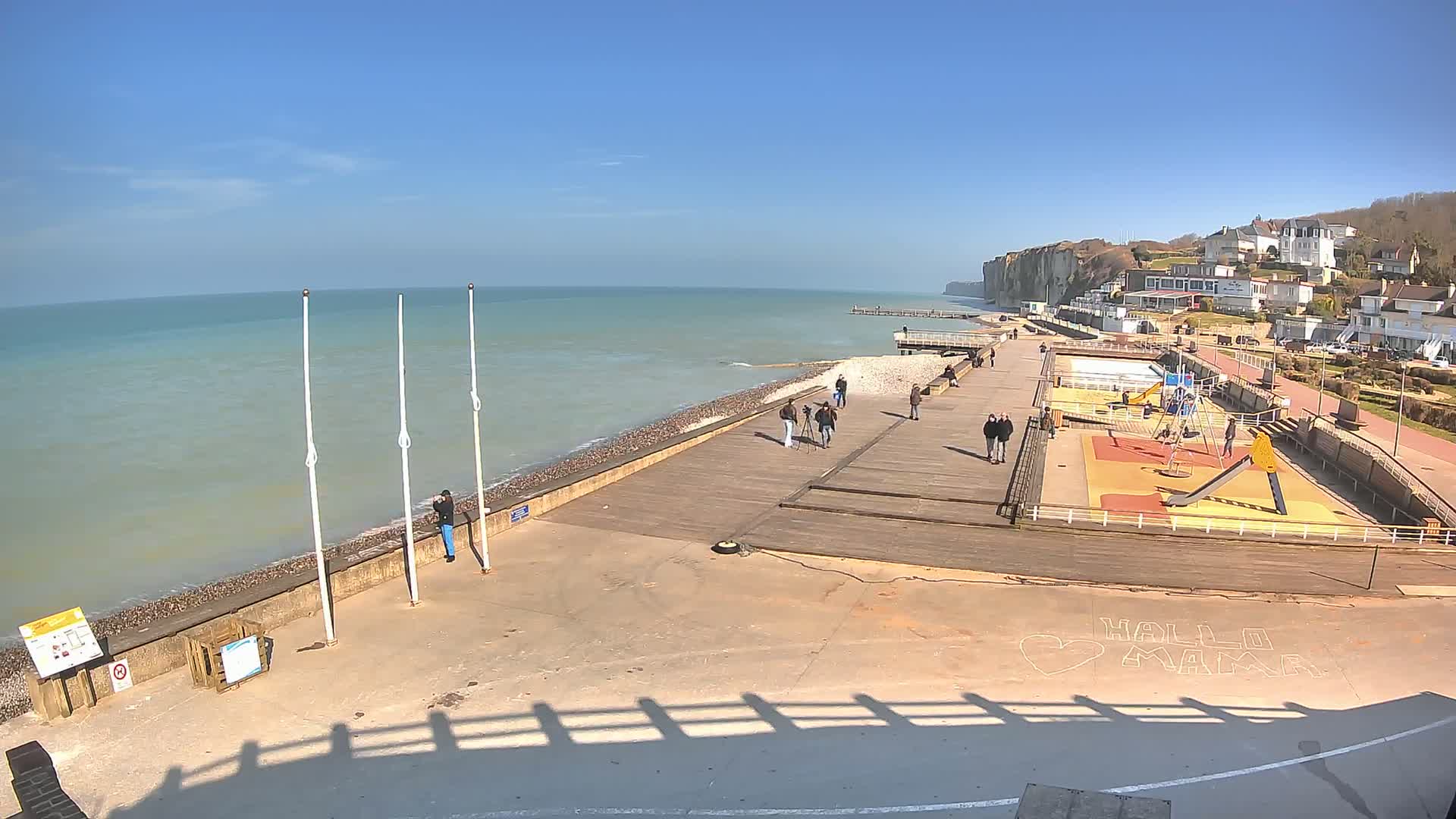 A sunny day at a seaside boardwalk shows a calm ocean, a pebble beach, a playground, and several people strolling along the wooden walkway near white cliffs in the distance.