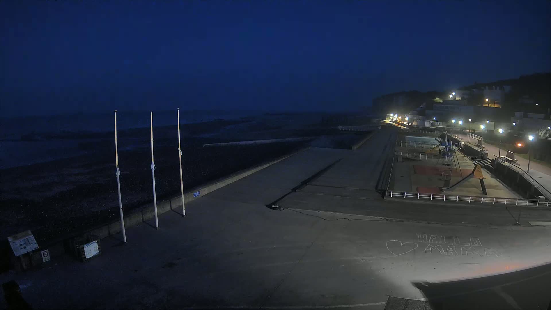 A dark, nighttime view of a seaside promenade with a playground and a mostly empty beach under a dusky sky.