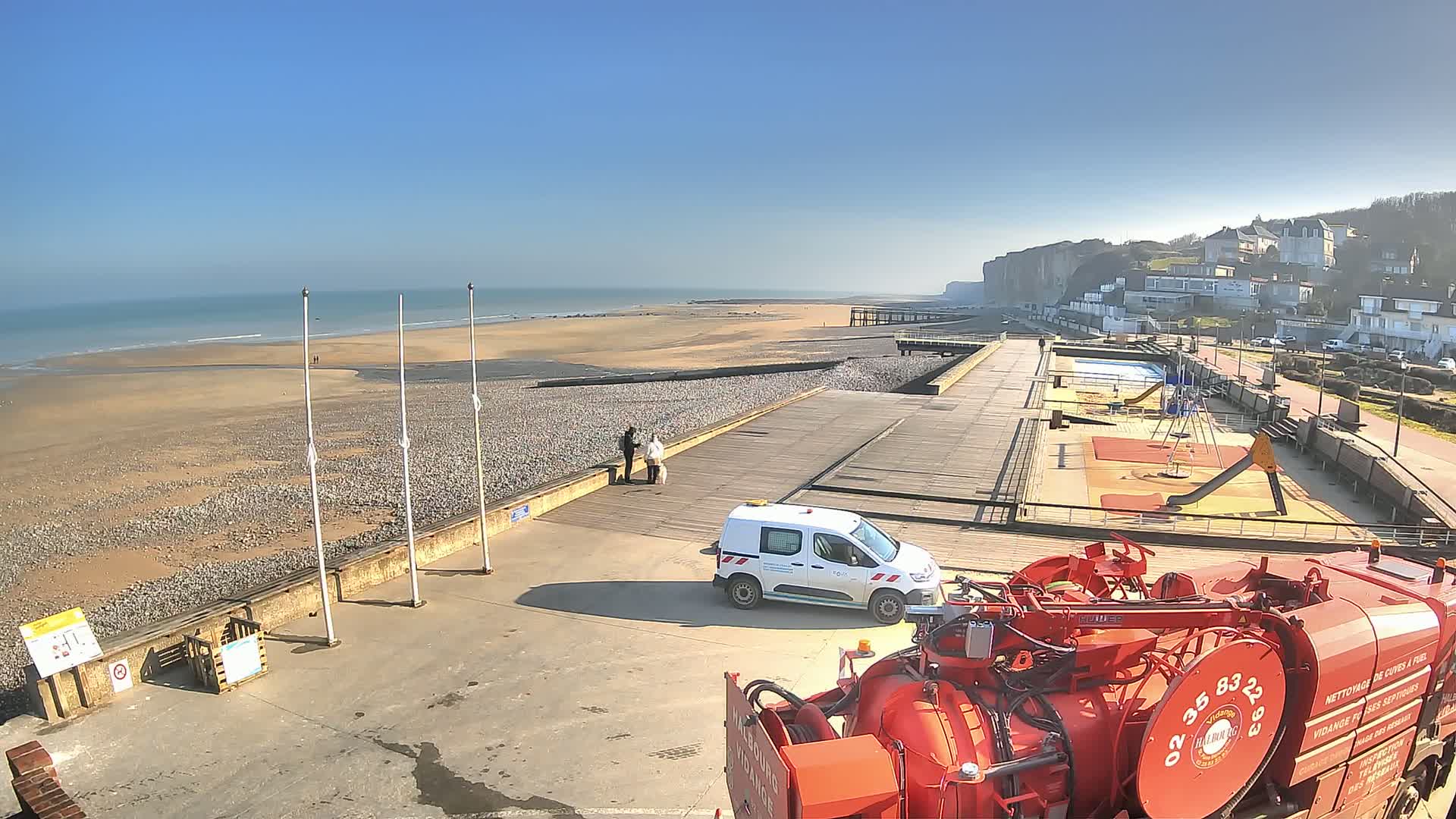 A seaside promenade on a sunny day features a sandy beach, a wooden boardwalk, a playground, a white van, and a large red sanitation truck.