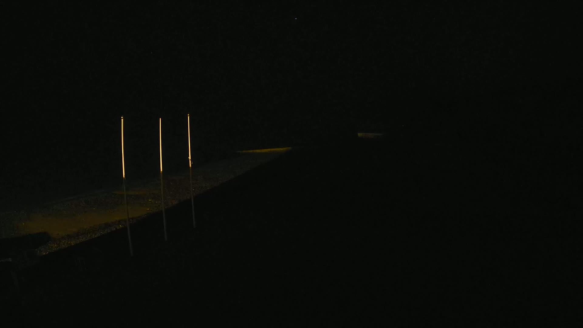 Three tall, thin lights illuminate a dark, flat ground at night.