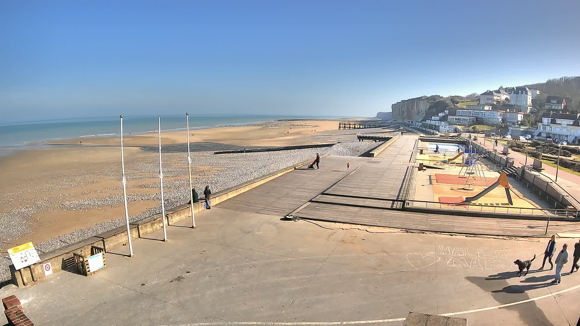 A sandy beach with a wooden boardwalk, playground, and a small town are visible under a clear blue sky.