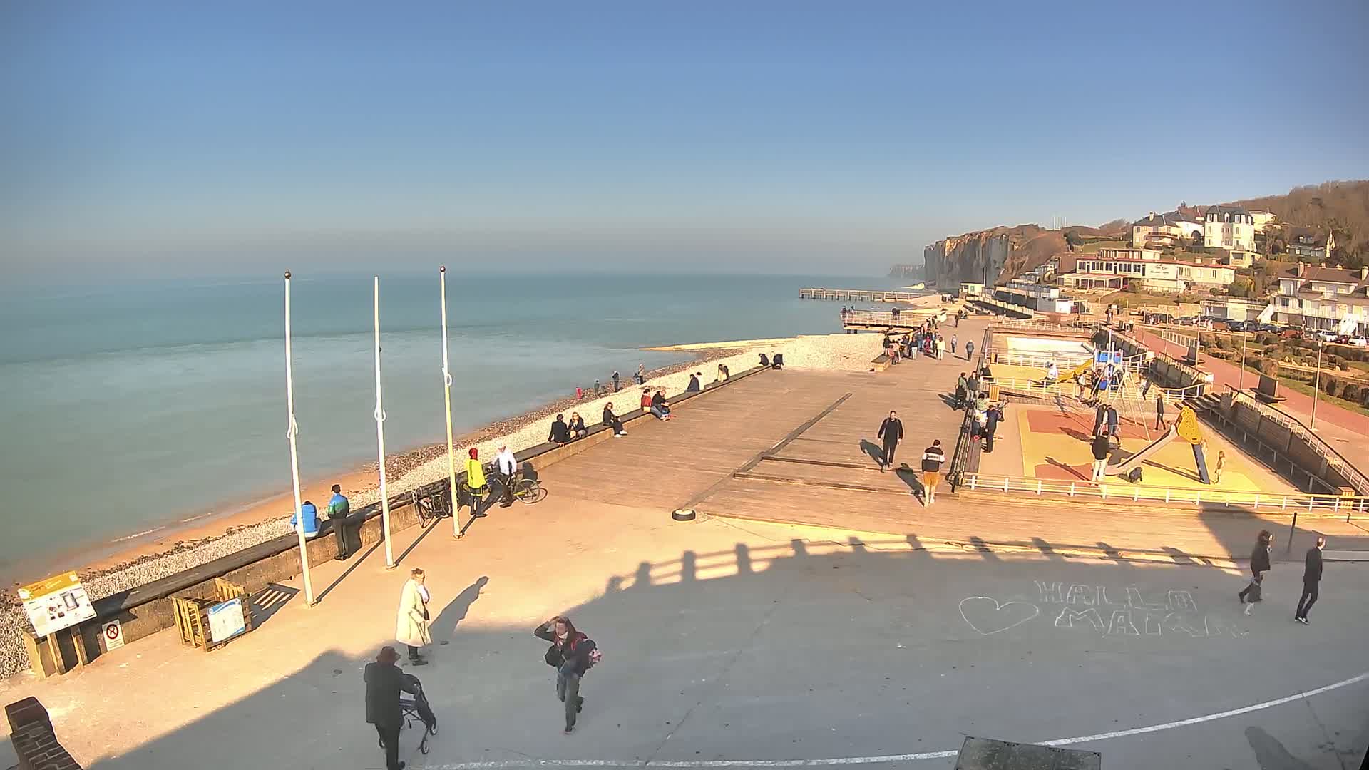 A sunny seaside town scene features a boardwalk with people walking, sitting, and playing on a playground, all overlooking a calm ocean and white cliffs in the distance.
