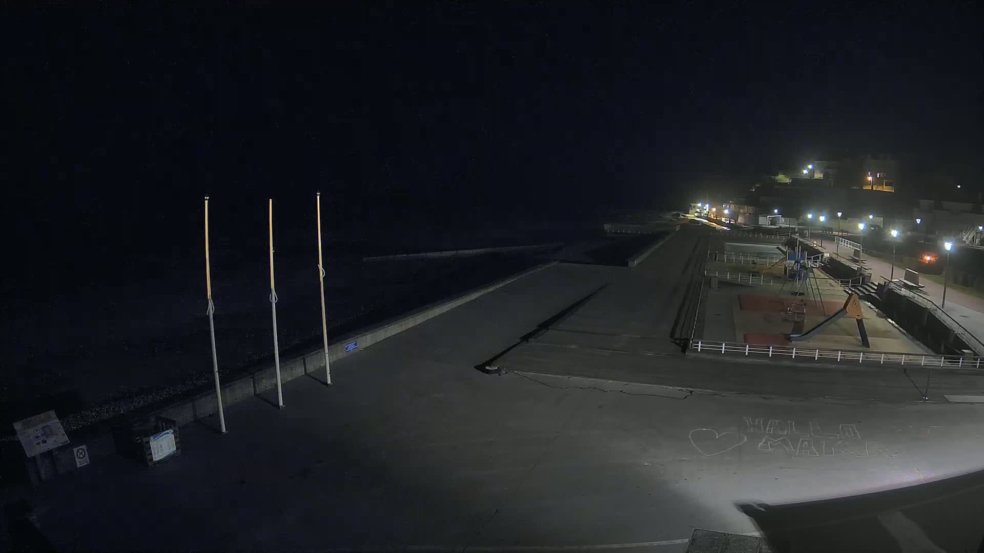 A nighttime view of a mostly empty seaside promenade with a playground and a small number of lights illuminating the scene.