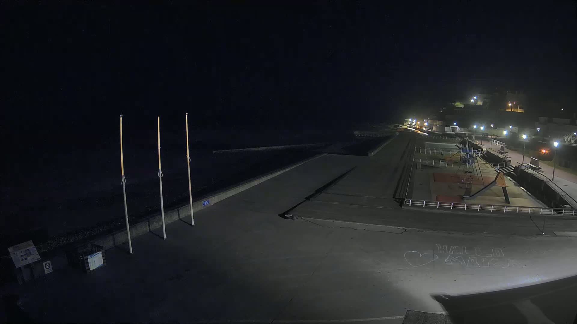 A nighttime view of an empty seaside promenade with a playground, lit by streetlights under a dark, clear sky.