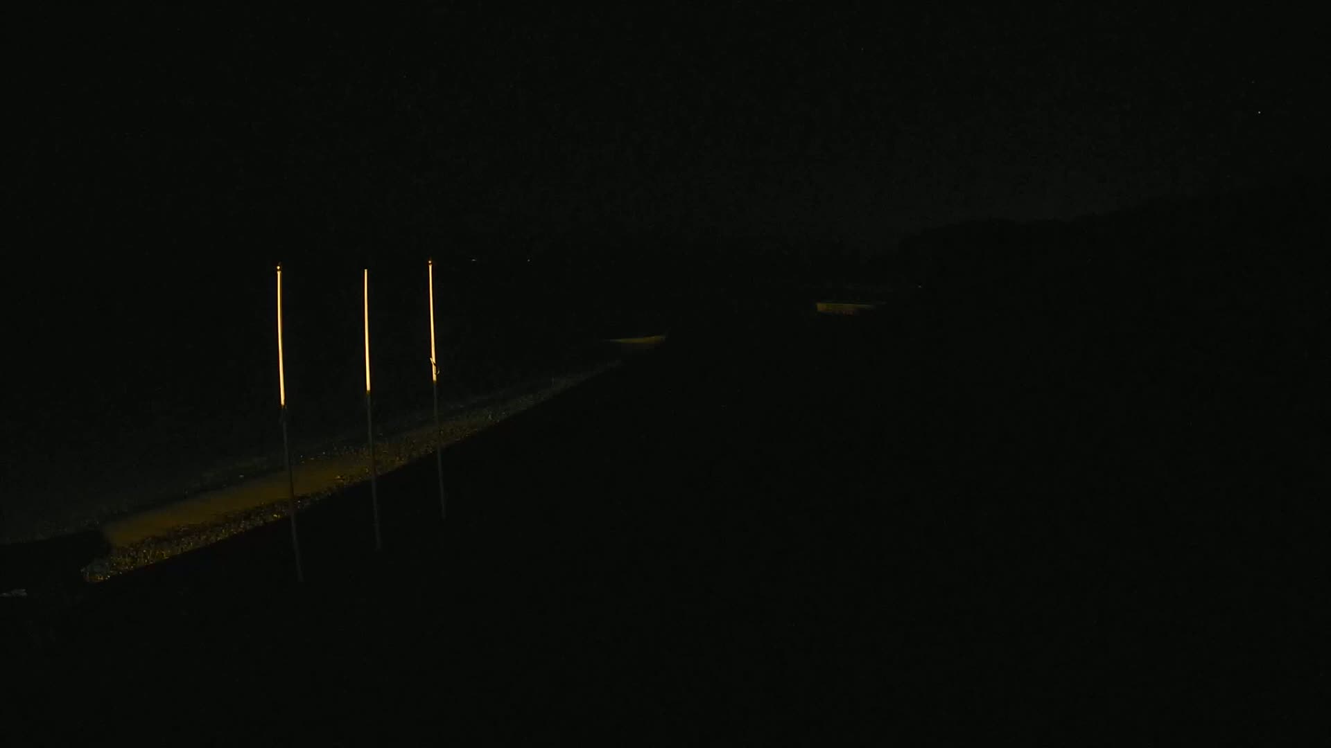 Three tall, thin, illuminated lights stand on a dark, paved surface at night.